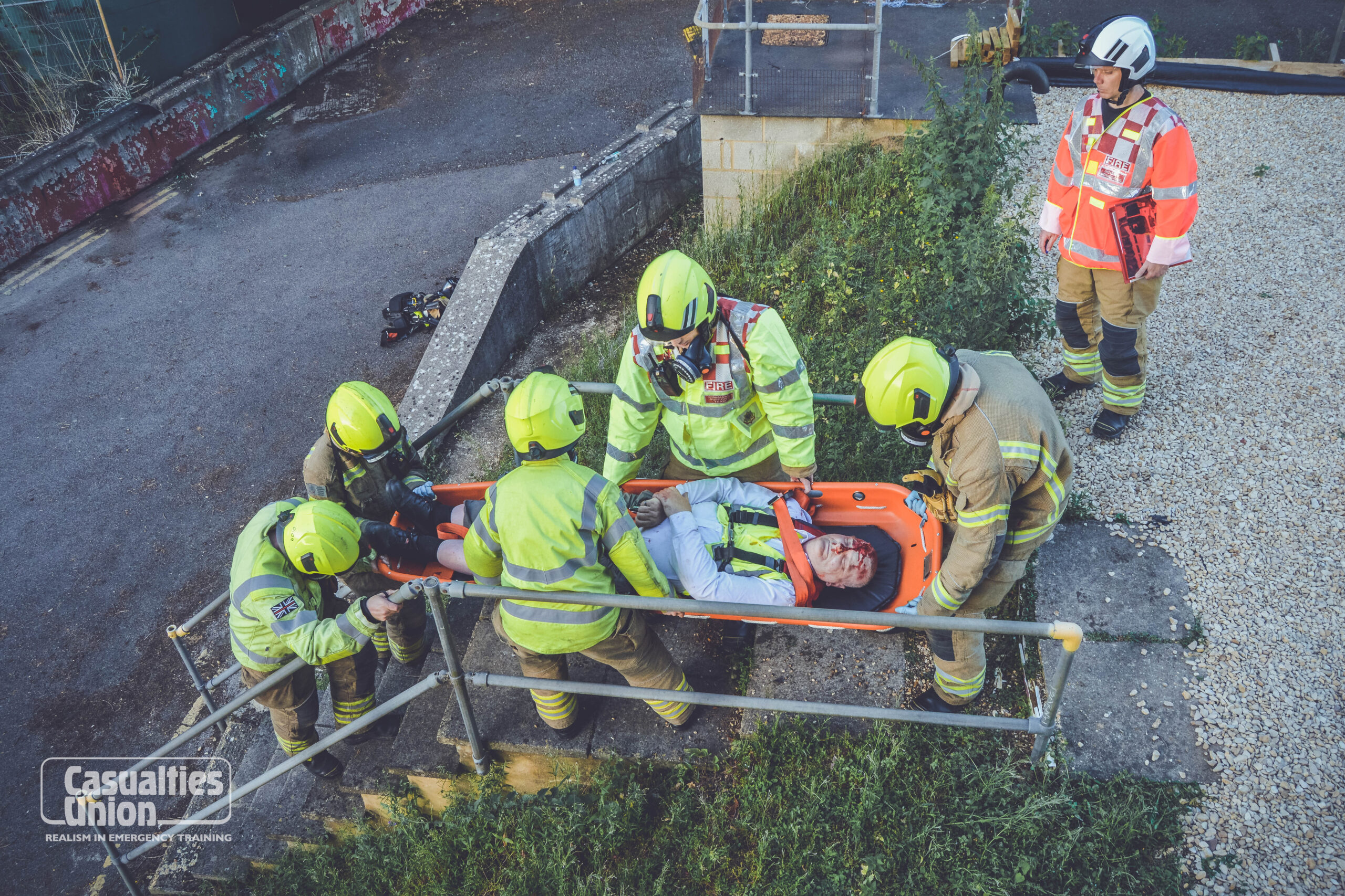 A person portraying someone who is injured (with a simulated head injury) is carried down stairs on a stretcher by a fire service